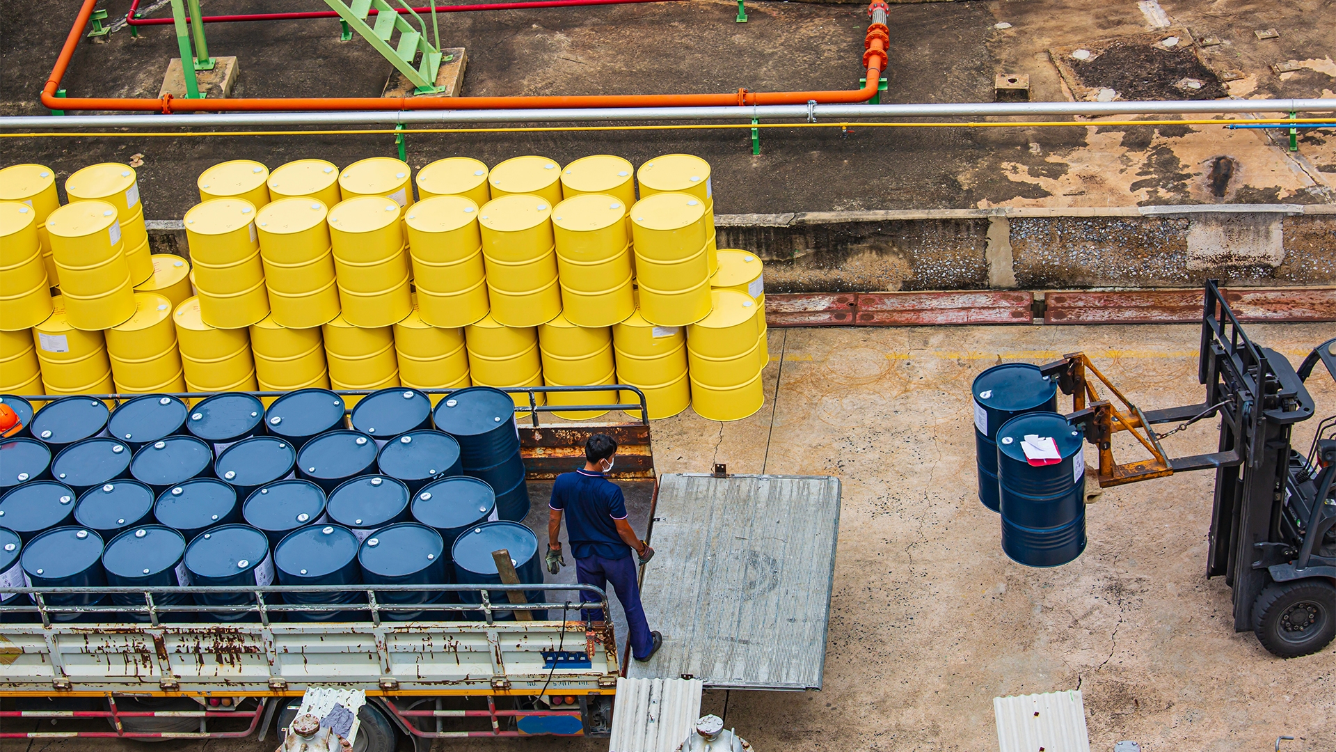 Lupa Ambiental | Vista aérea de tambores industriais amarelos empilhados e azuis sendo carregados em um caminhão por um funcionário e uma empilhadeira em pátio de fábrica.